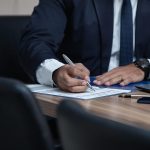 Homme en costume noir écrivant sur une feuille à une table en bois